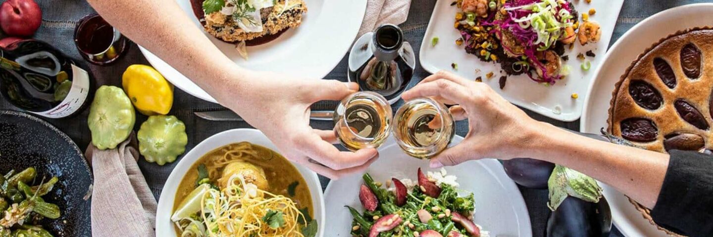 Friends toast wine glasses together above a table filled with food deaturing local, seasonal ingredients from Willamette Valley.