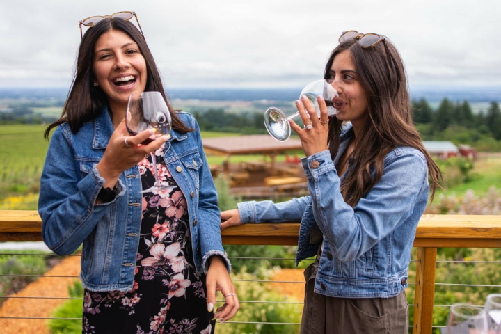 Two young women sipping wine and laughing on the deck of a winery overlooking the vineyard below. Both women are wearing summer dresses with denim jean jackets and sunglasses on top of their heads. 