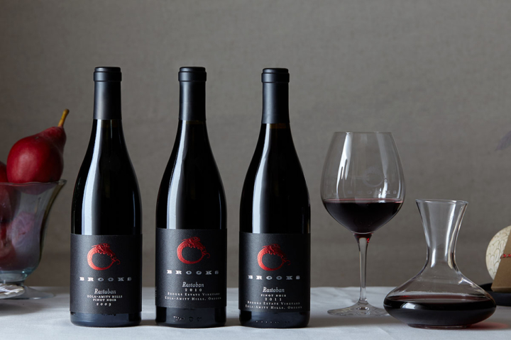 A studio image of three wine bottles with black labels and a wine glass and glass decanter filled with Pinot Noir, the most versatile wine for cooking and pairing. 