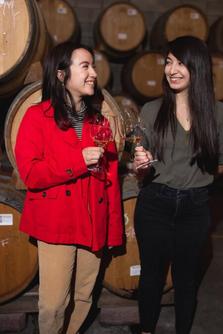 Two friends smile and toast their wine glasses together while touring the barrel room of a winery. 