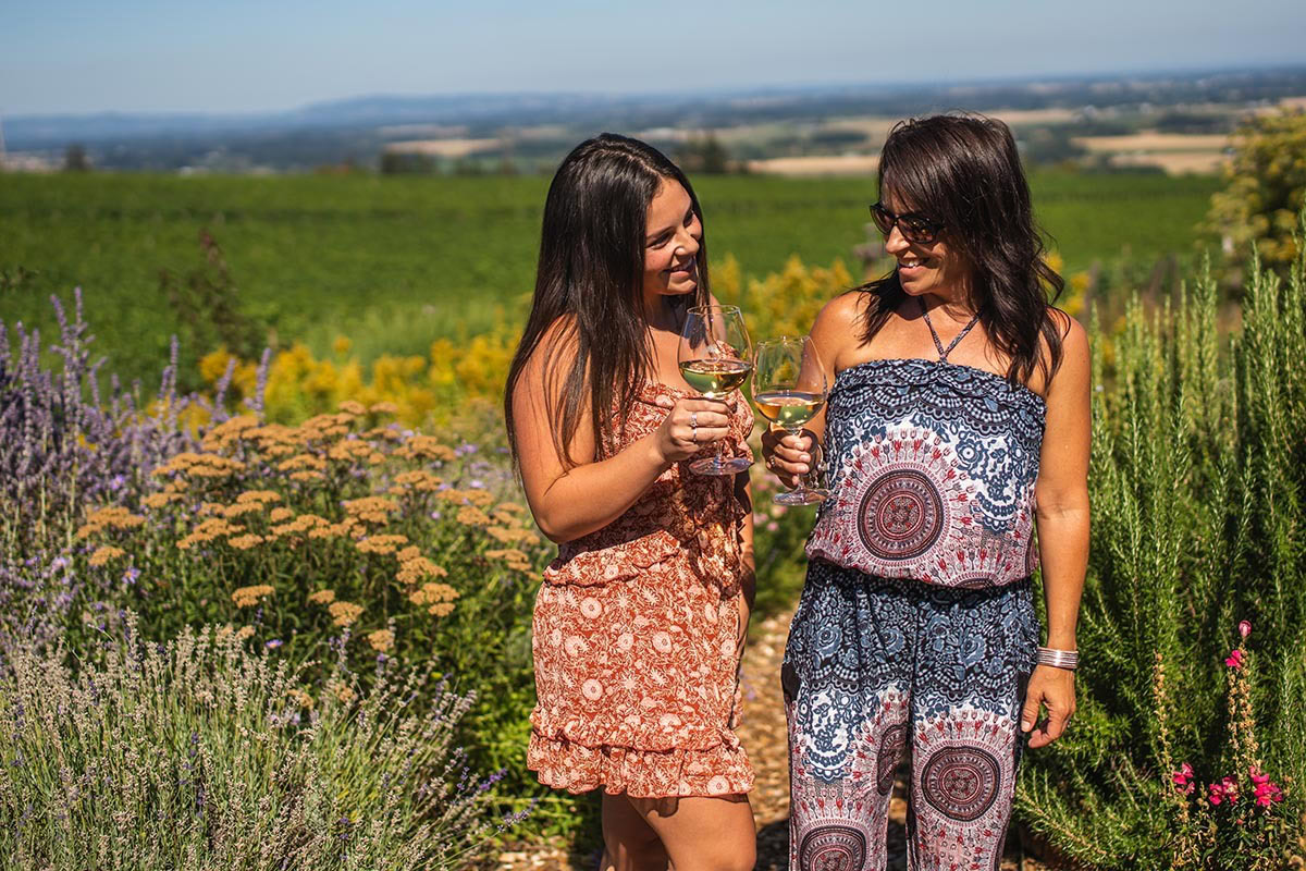 Two young women pose for an Instagram photo while tasting wine and walking through the lush garden at Brooks Wine on a sunny day.