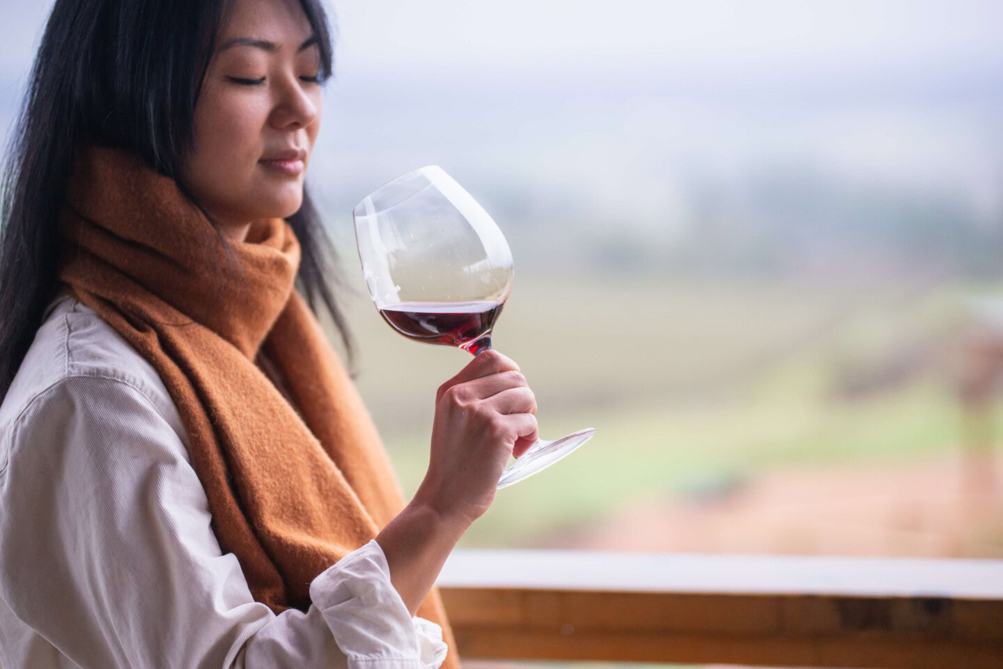 Asian woman tastes Pinot Noir from Willamette Valley while wearing a mustard yellow, cashmere scarf and white, cotton, long sleeve shirt with the sleeves rolled to her elbows.