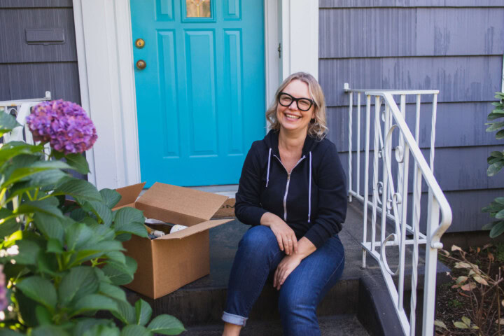 A blonde woman sits on the front stoop of her doorstep in front of a bright blue door. She is wearing a black zip up hoodie and dark jeans. She is smiling and sitting next to a wine delivery.