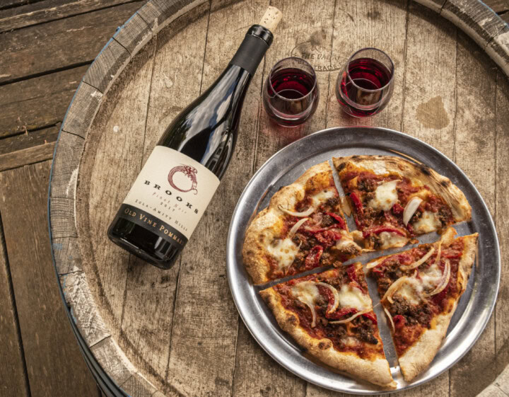 Using an old wine barrel as a table, a bottle of earthy pinot noir from Brooks Wine in Willamette Valley Oregon is served alongside a freshly baked pizza of sausage and rich tomato sauce. 