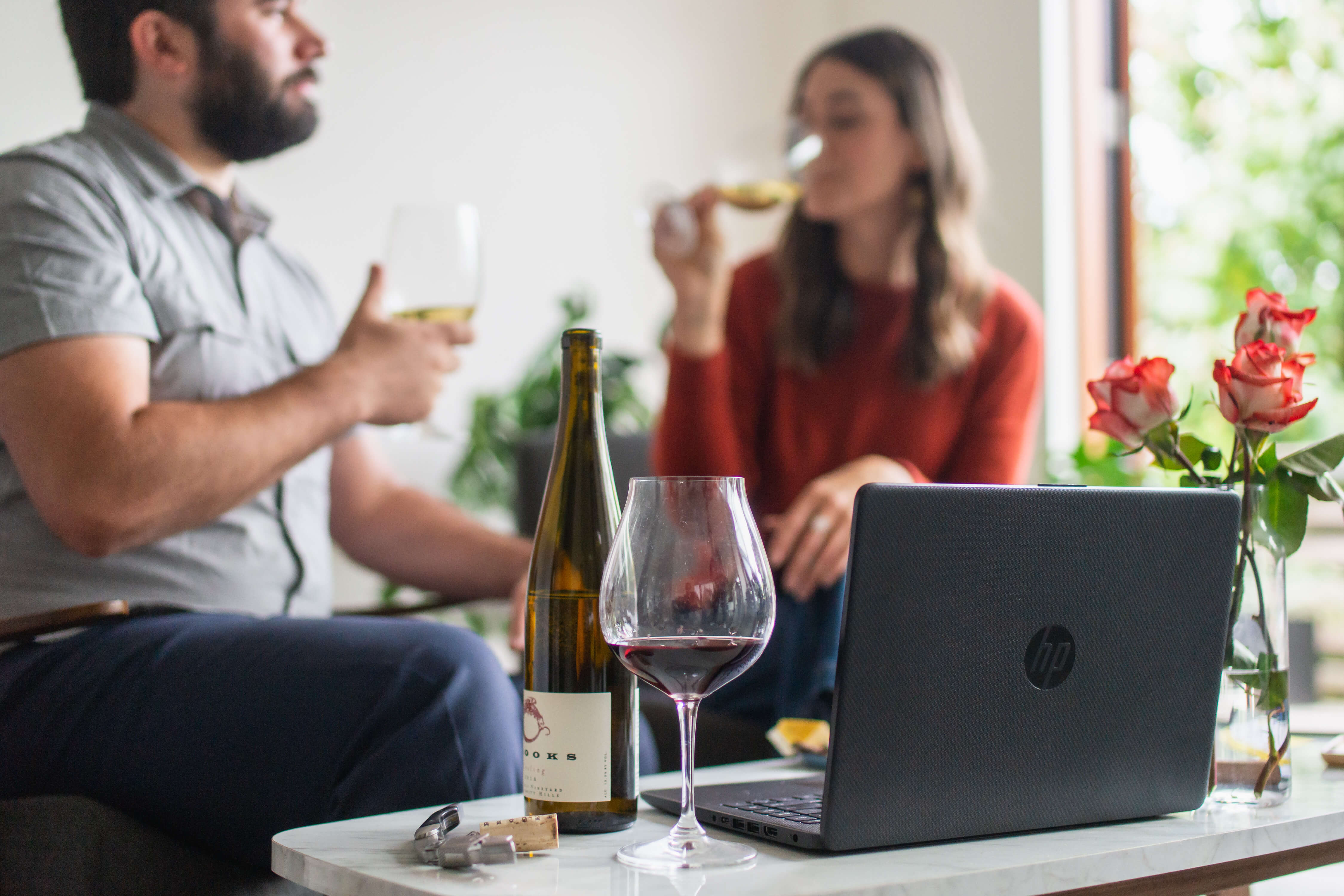 A man and a woman sit together at home while tasting and ordering wine online with their laptop. She is wearing a red long sleeve shirt and he is wearing a light gray short sleeve polo shirt with jeans.