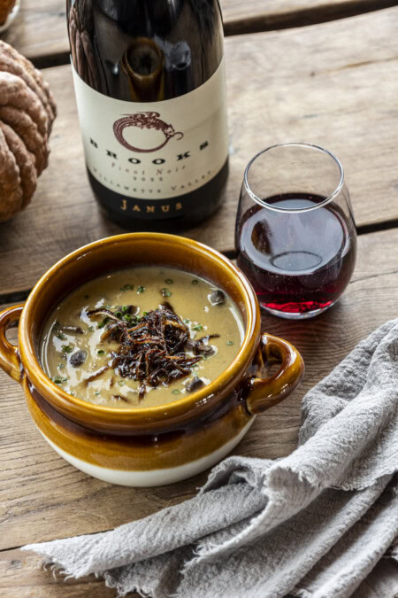 A wooden table features a rustic, gray napkin beside a cozy bowl of pumpkin and mushroom soup paired with a red-fruit focused Pinot Noir from Willamette Valley. 