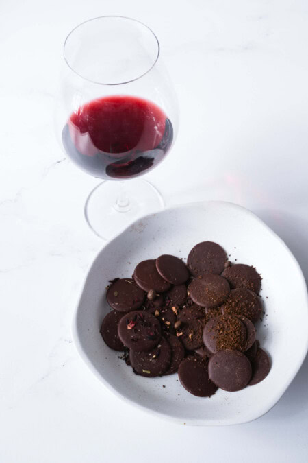 A white marble counter showcases a white, asymmetrical bowl is filled with round disks of dark chocolate while a glass of red wine rests beside it. 