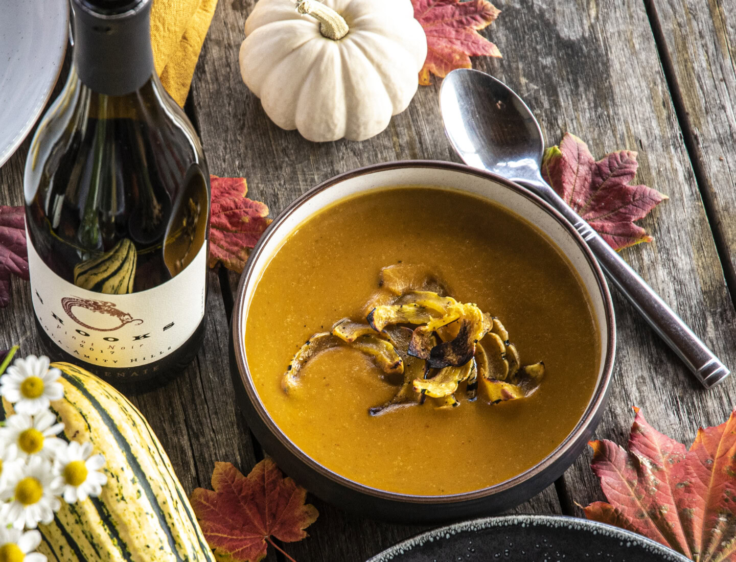 A table decorated with various autumn squash, orange flowers, and a bottle of Brooks Pinot Noir surround a bowl of pumpkin soup with roasted delicata squash on top.