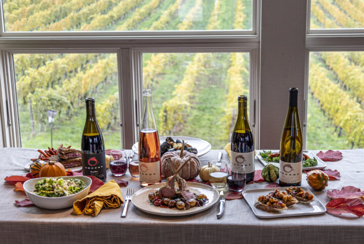 A Friendsgiving feast with a variety of dishes and four bottles of wine sit on a gray linen tablecloth decorated with pumpkins and red leaves with a grape vineyard prominently featured outside the large windows in the background. 