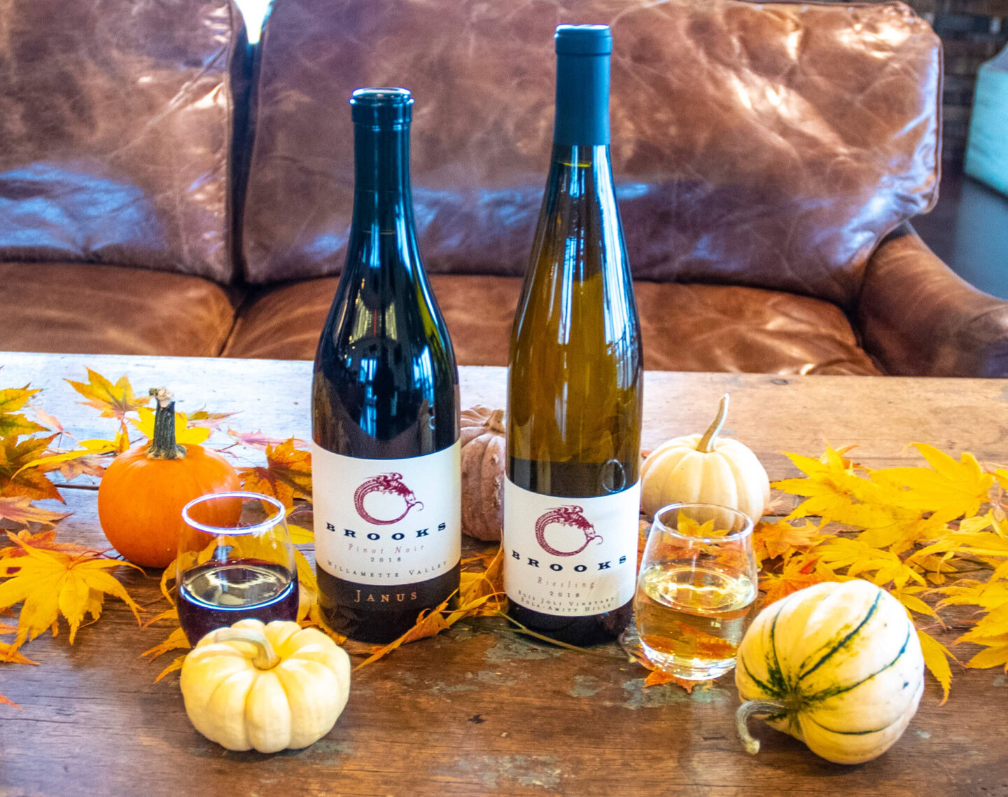 A bottle of Riesling and a bottle of Pinot Noir res in a wooden table decorated yellow maple leaves and small white and orange pumpkins with a brown leather couch in the background for guests to sit on during Friendsgiving.