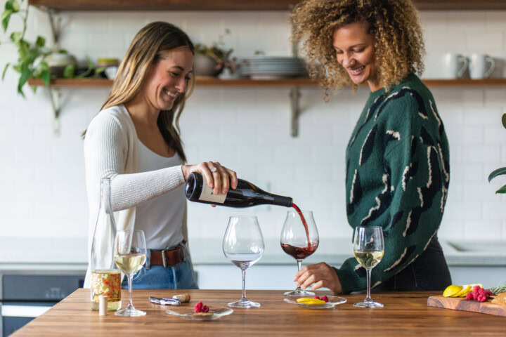 A young woman with blonde hair and white sweater pours a glass of Pinot Noir for a young woman with dark textured hair in a green sweater. 