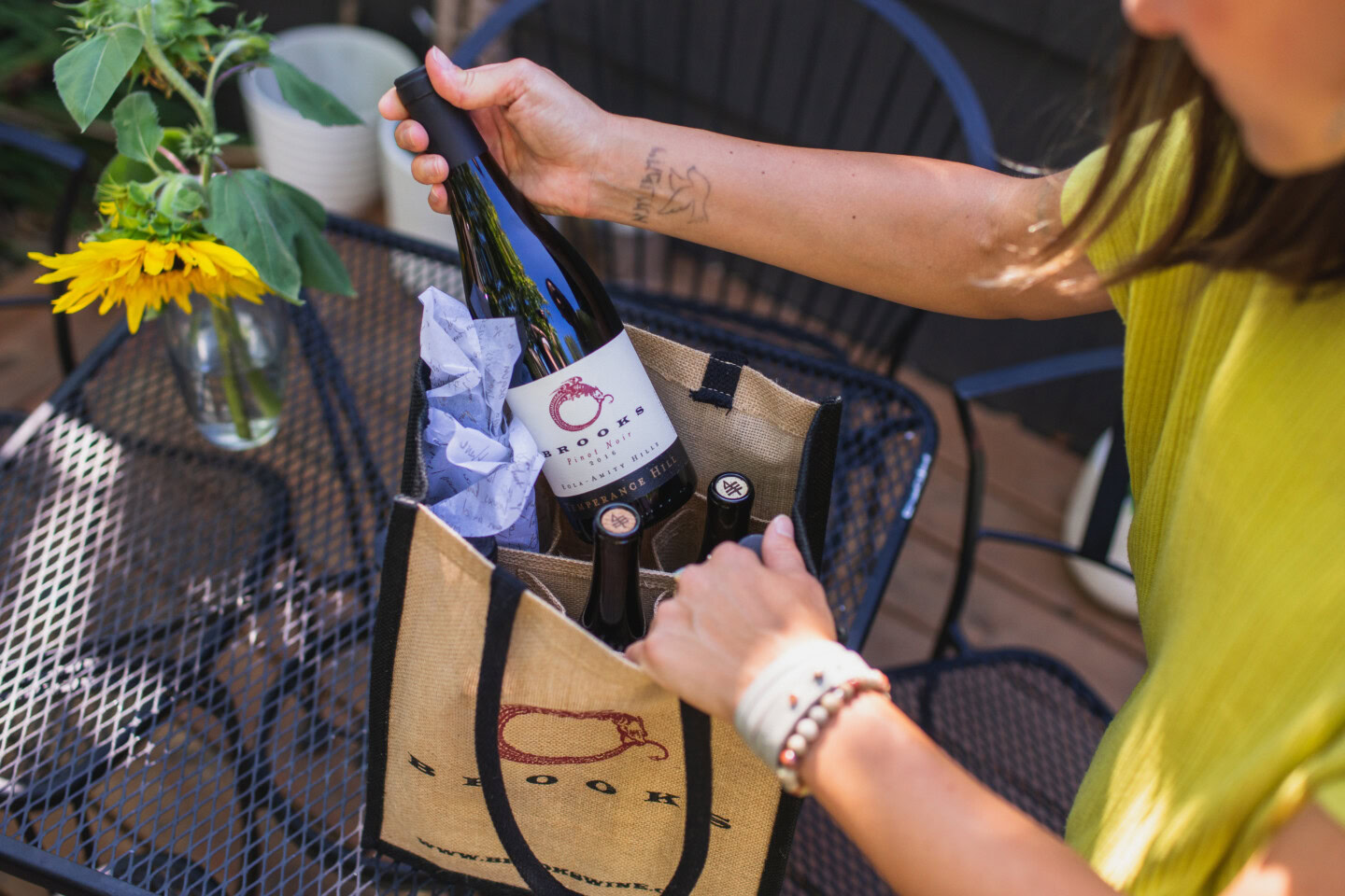 A woman in a yellow blouse examines a bottle of wine from a gift bag delivered to her home's door step.