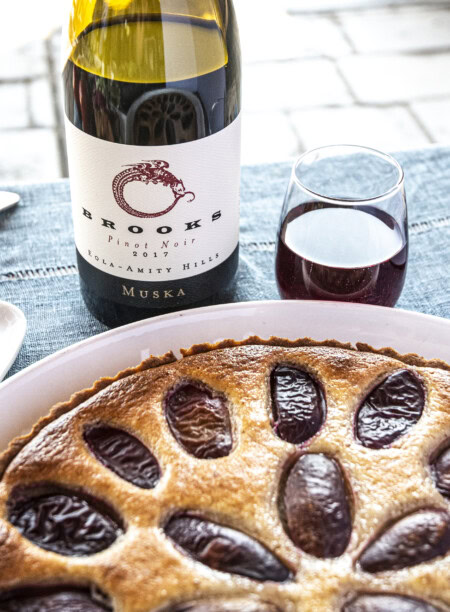 A bottle of Pinot Noir rests on a blue linen tablecloth next to a freshly baked plum cake. 