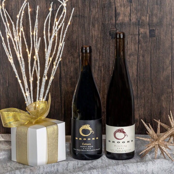 A bottle of Pinot Noir and a bottle of Riesling are set in front of a rustic wooden backdrop with golden star tree decorations on the right-hand side and the left-hand side includes a white gift box with golden ribbon and Birch branches decorated with twinkly lights. 