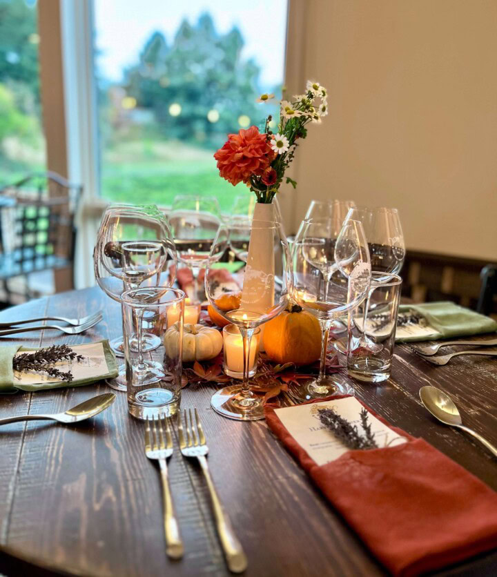 A wooden table is set for entertaining dinner guests with dark orange cloth napkins folded to hold a menu and sprig of lavender with the center of the table displaying votive candles, dried leaves, small pumpkins, and a thin white vase holding orange flowers. 