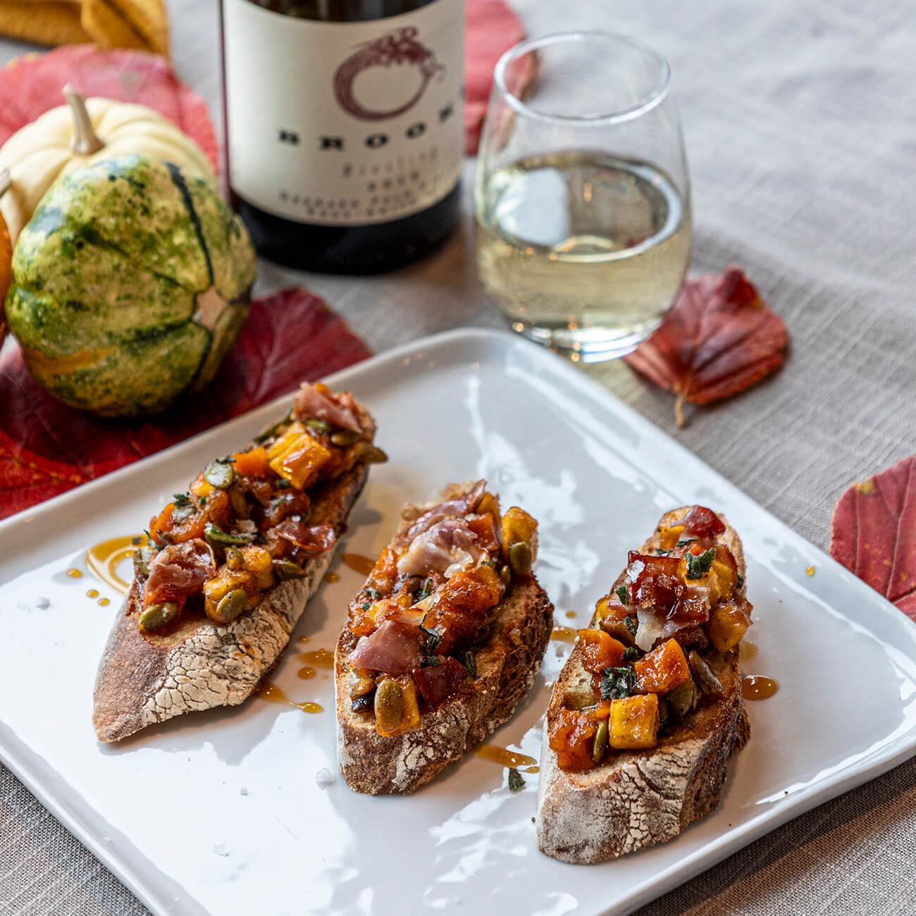 A crisp glass of white wine paired with an appetizer of squash and pecan bruschetta on a table decorated with a yellow gourd.