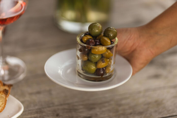 A server's hand carries a small white plate supporting a small glass bowl of mixed olives as an appetizer to pair with wine. 