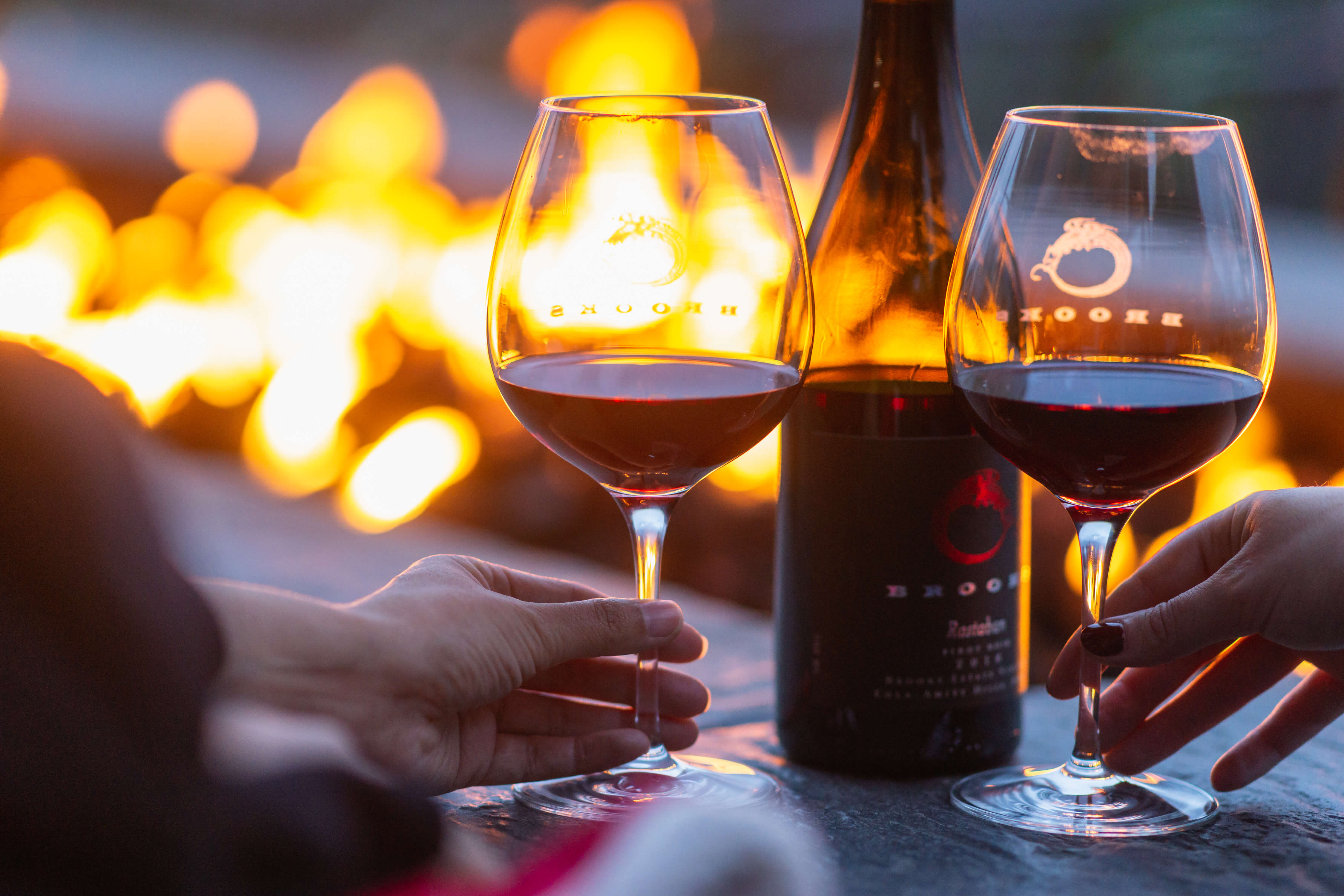 Two sets of hands rest on glasses of Pinot Noir with a wine bottle between then and the flames from a warm fire pit in the near distance.