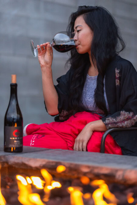 A young Asian woman with long black hair sits next to a cozy fire pit and takes a sip of a glass of red wine while wrapped in a fluffy, red, sherpa blanket. 