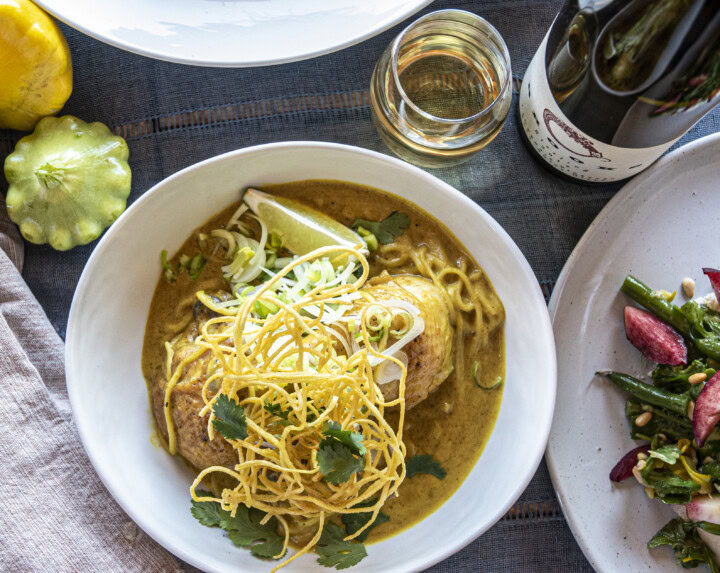 A cozy bowl of Thai chicken curry and crunchy noodles rests on a table covered in a blue linen table cloth beside a glass of Riesling, an asparagus and radish salad, and squash as table decor. 