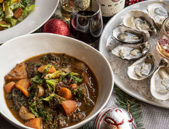 A close up view of a warm, cozy dinner featuring beef stew, oysters on ice, hearty Caesar salad, and red wine alongside tree ornaments and sprigs of pine tree as table decorations. 