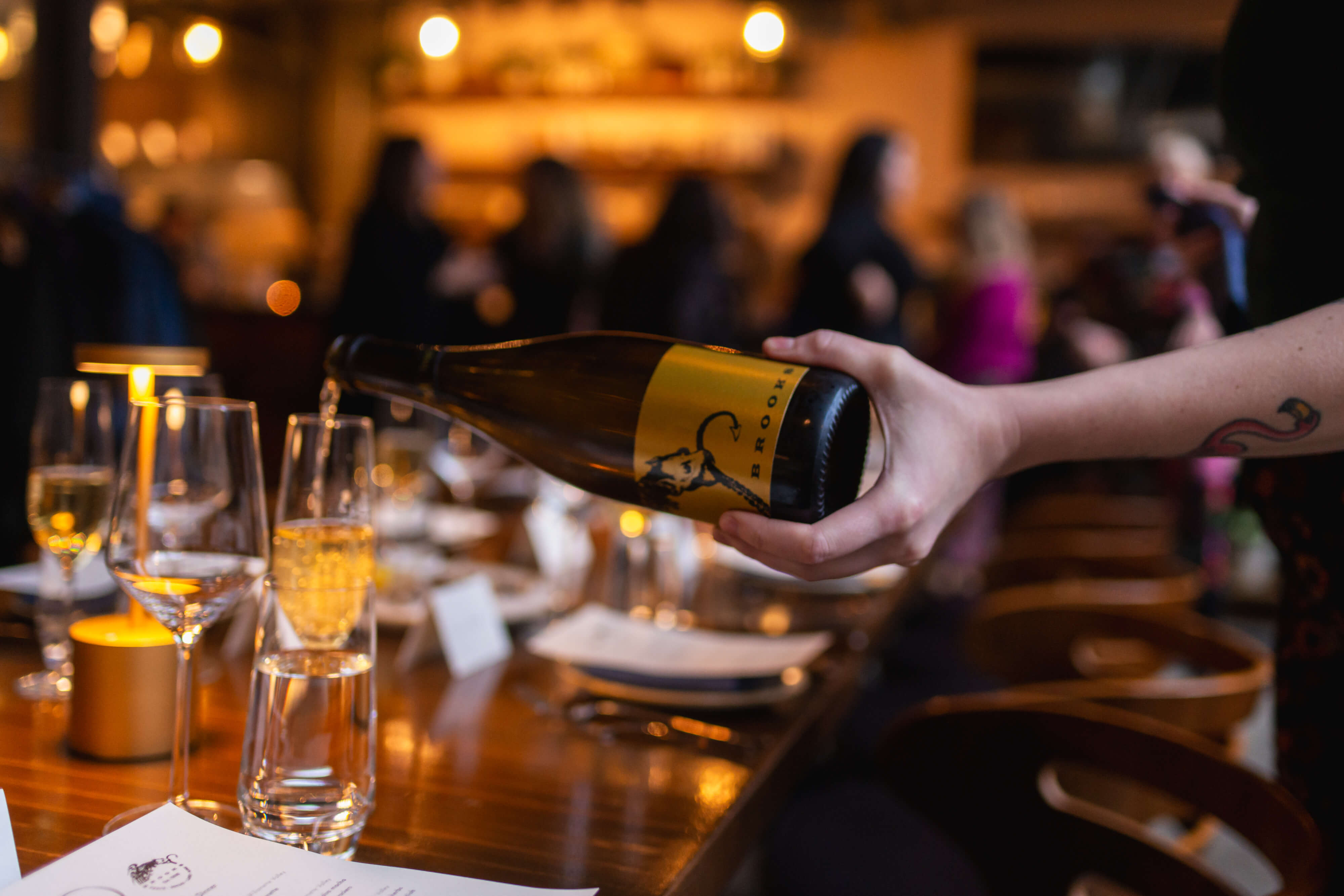 A glass of sparkling wine is being poured on a long table set for a cozy winter dinner party inside t room with dim lighting.