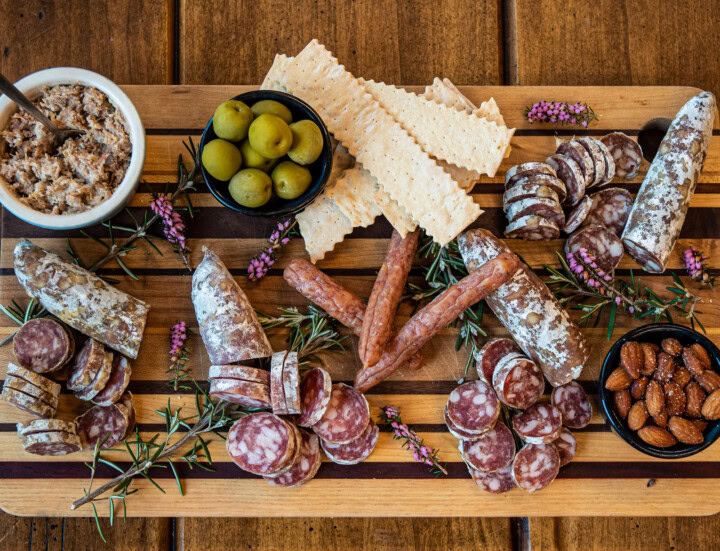 An overhead shot of a striped wooden serving board displaying an appetizer of six different kinds of charcuterie as well as salted almonds, green olives, long crackers, and decorative sprigs of lavendar. 