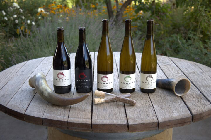 A selection of 100% biodynamic Pinot Noir and Riesling from Brooks Wine in Oregon sit on a circular wooden table with a plus garden in the background. 