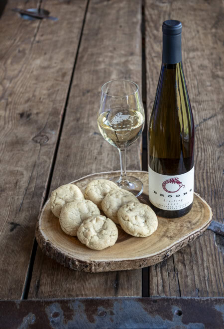 A rustic wooden table is set with a medium dry Riesling is paired with six white chocolate chip cookies served on a board made from a cross-section of a tree. 