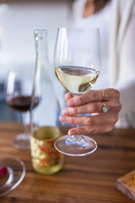 A woman in a white long sleeve shirt offers a glass of white wine toward the camera. 