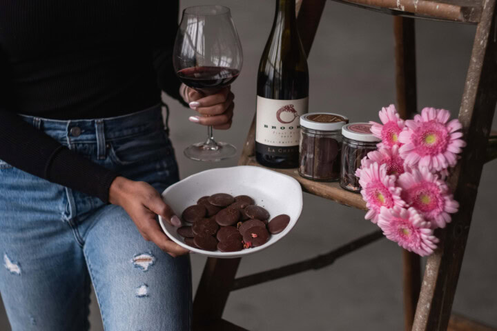 A woman wearing a black, long sleeved shirt and denim jeans holds a white bowl filled with dark chocolate disks and a glass of red wine while a wooden ladder is used to rest a bottle of Pinot Noir, two jars of chocolate disks, and a bouquet of pink flowers. 