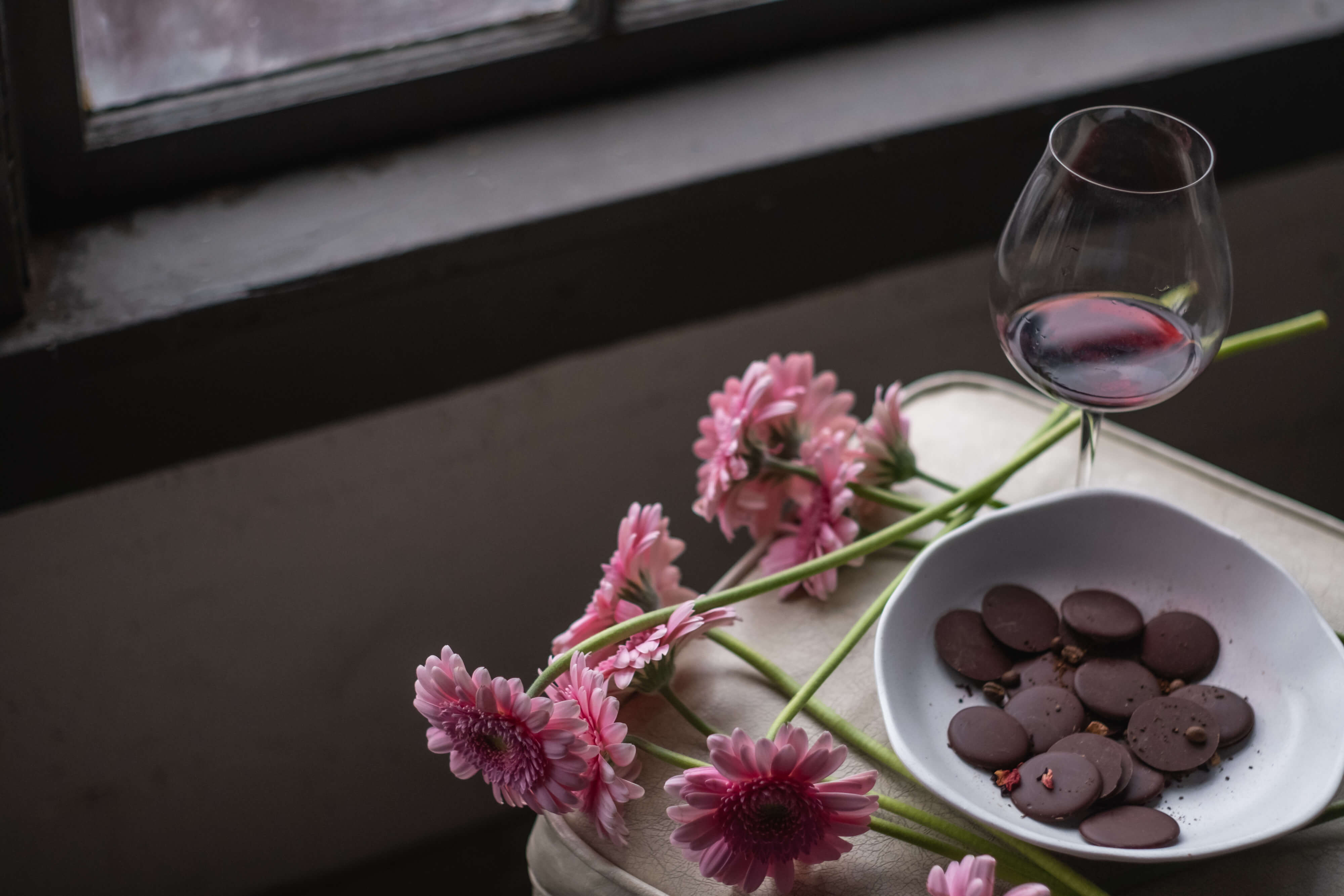 A white bowl of dark chocolate disks rests on a wooden stool next to a window with pink flowers set on the side of the bowl as well as a glass of red wine.