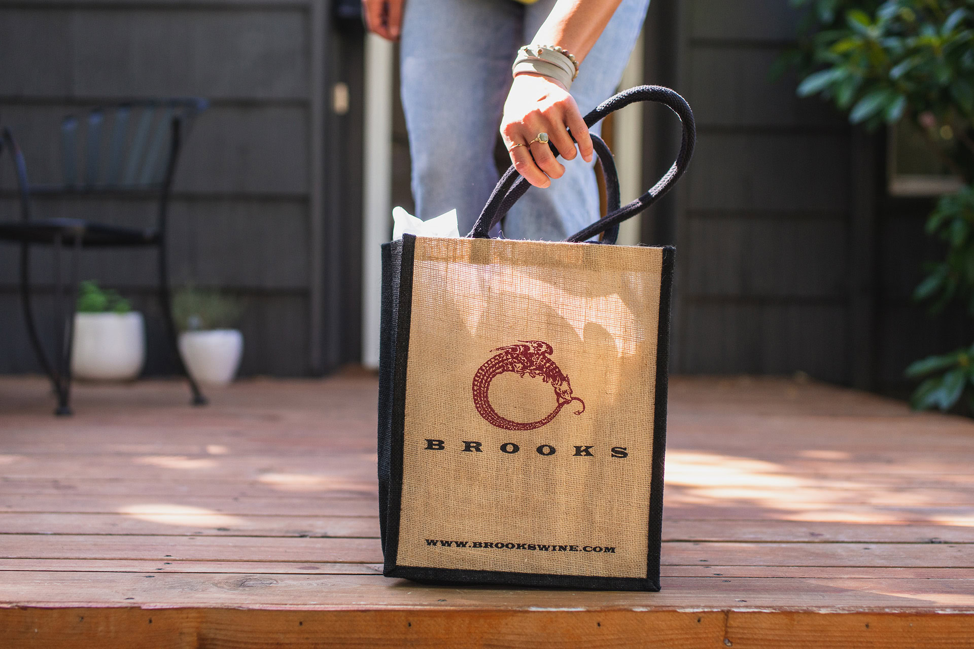 Jute Wine Bag featuring Brooks Logo. Woman in collecting bag from her front porch to take into house.