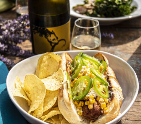 A photo of a hot dog topped with elote corn and jalapeno slices and paired with tortilla chips and sparkling Riesling for a very fun pairing on Super Bowl Sunday! 