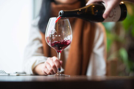 A woman in a white shirt and yellow scarf sits in the background of a wine glass filling with a heavy pour of Pinot Noir ordered to her preferences. 