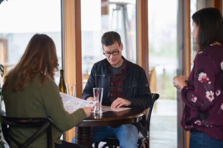 A man with black thick rimmed glasses, dark jacket, and denim jeans reviews the wine list and discussing his order with the hostess. 