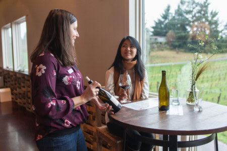 A young woman with dark hair and a yellow scarf sits at a round wooden table next to a large window overlooking a vineyard while the hostess describes the wine she has ordered. 