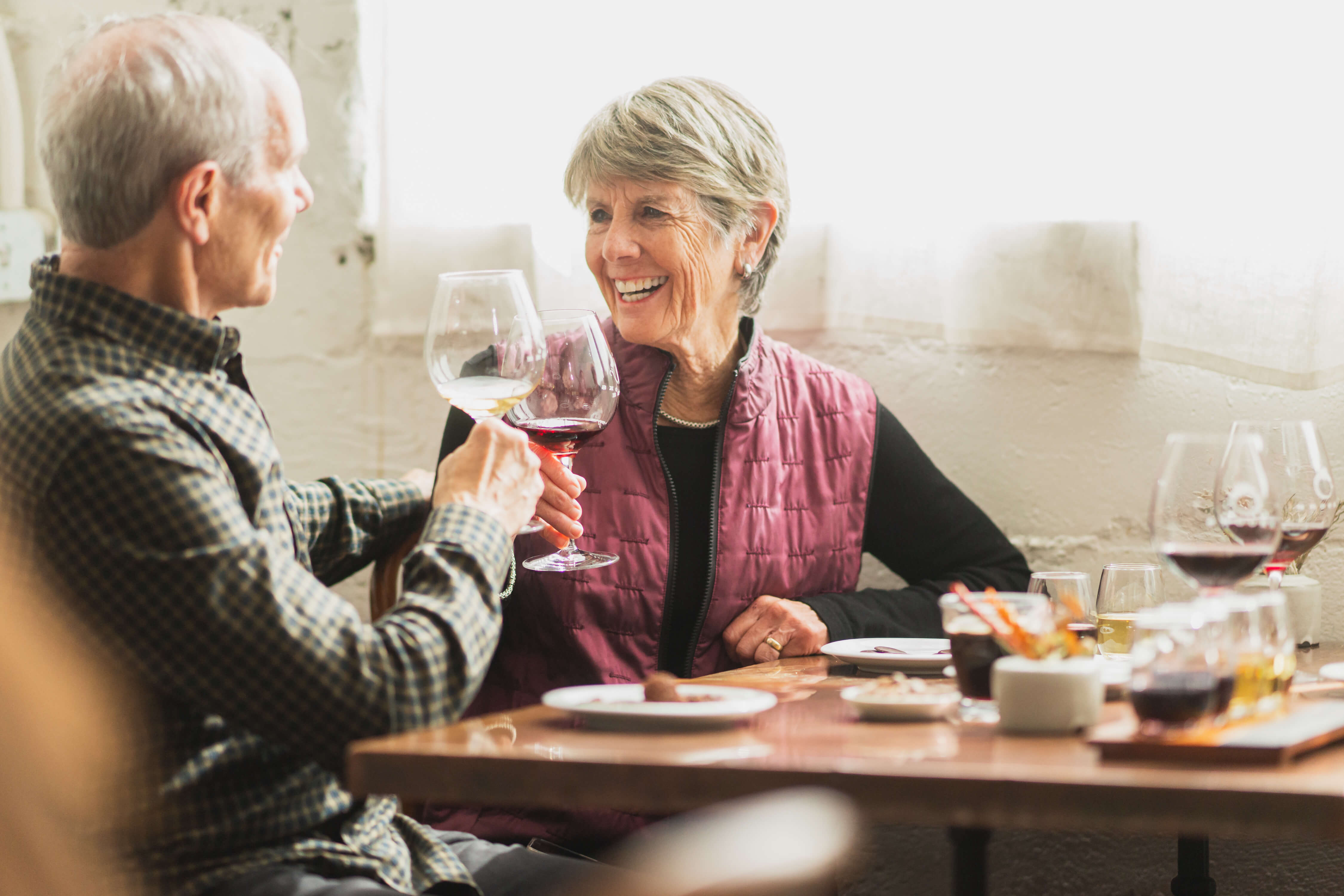 A mature man in a green sweater and mature woman in a pink vest sit smiling at one another while touching their wine glasses in a toast after a successful order.