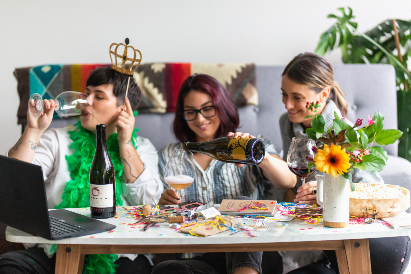 Three friends are draped with party favors as they pour and sip wine in front of a screen as they prepare to cheer on their football team in the Super Bowl.