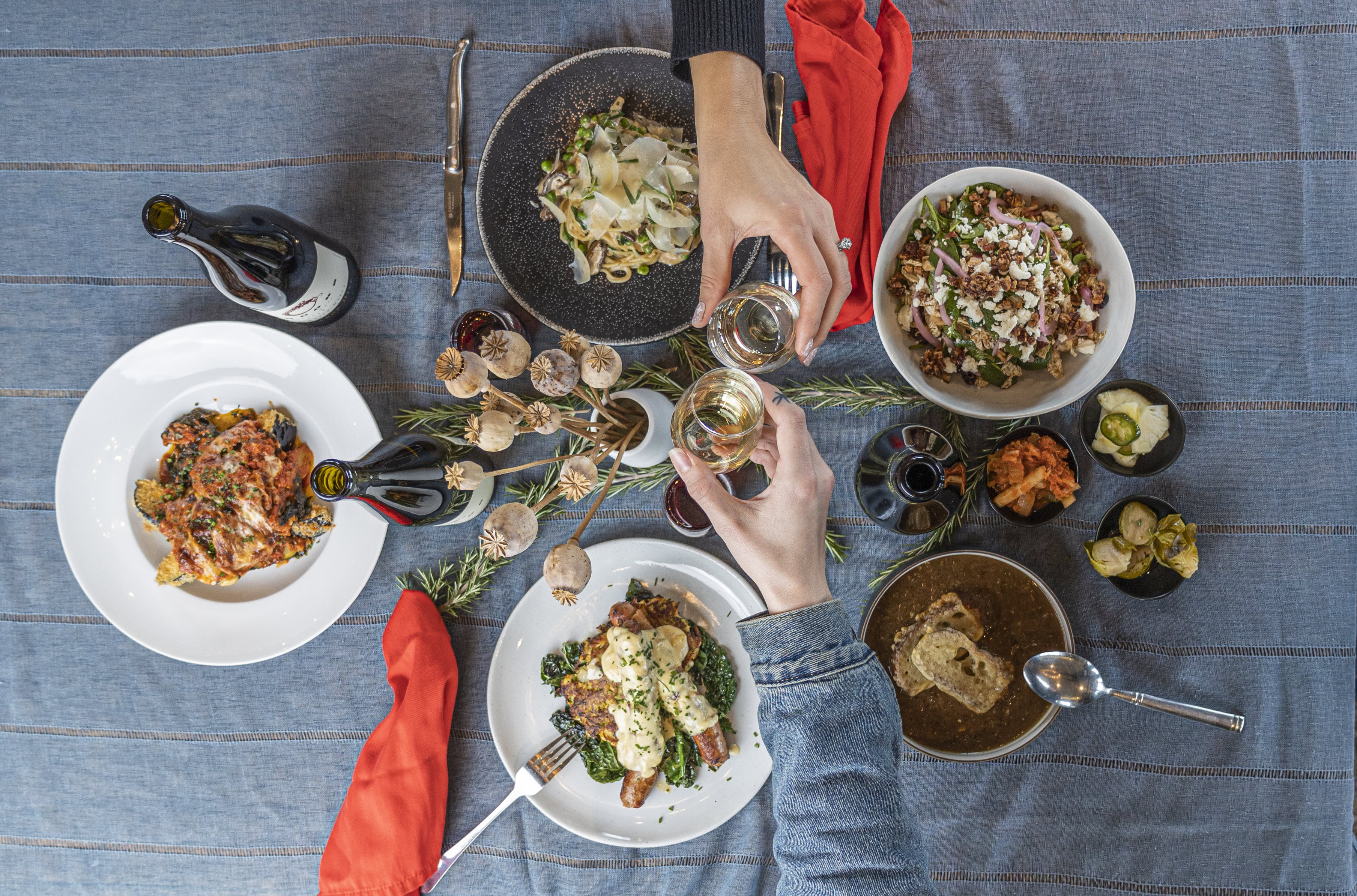 An overhead shot of a table draped in a blue tablecloth with bright orange cloth napkins and four dishes each cooked with wine as two diners extend their wine glasses across the table in a toast.