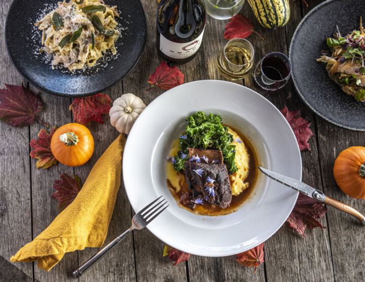 A rustic wooden table prominently displays multiple glasses of wine and a white bowl filled with yellow polenta, kale, and red wine-braised short ribs as well as a black plate carrying thick noodles cooked in white wine with cheese and fried sage leaves. 