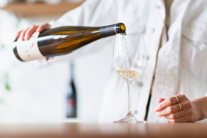 A woman in a white denim jacket joyfully pours a glass of Riesling wine into a slender wine glass from her latest wine buying adventure. 