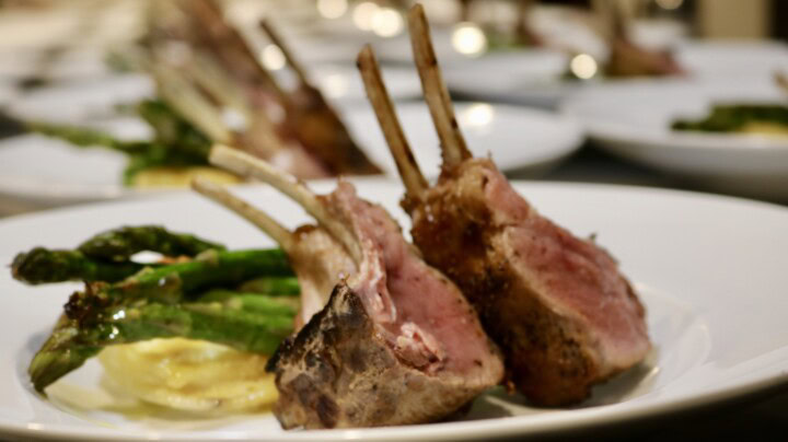 Rows of plates prepared for a dinner each hold racks of lamb resting on sliced potatoes and green asparagus for a pairing with Pinot Noir wine. 